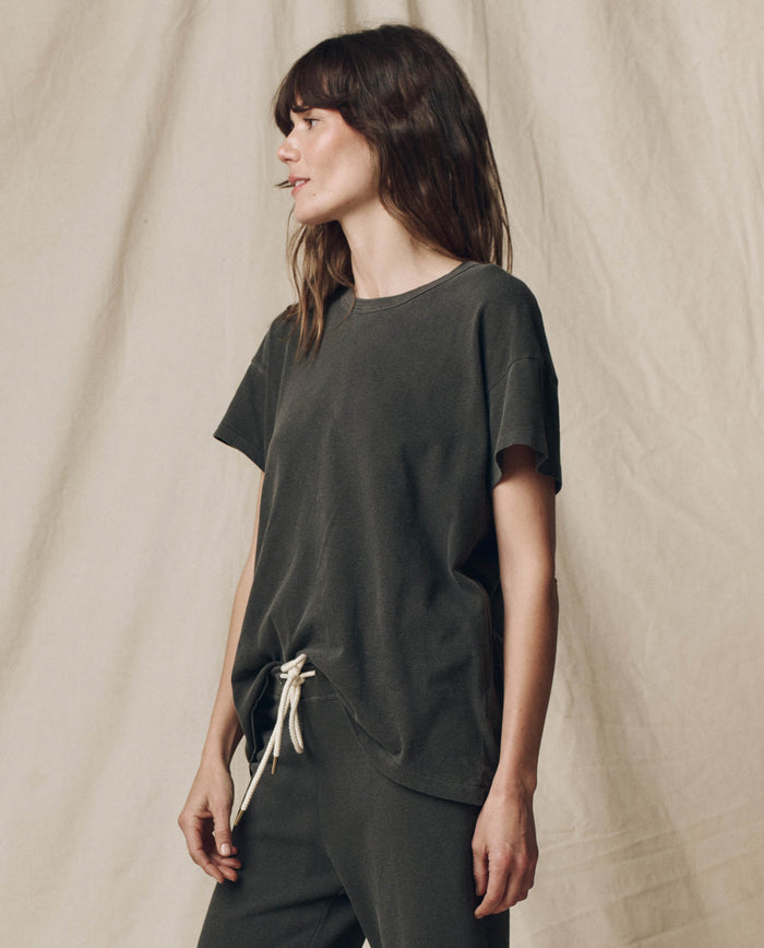 A woman with long hair stands sideways, wearing THE GREAT. CORE KNITS' The Boxy Crew. Solid in Washed Black, a 100% cotton t-shirt, and light-colored drawstring pants. A neutral fabric backdrop complements her timeless U.S.A.-made attire.
