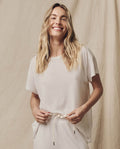 A person with long blonde hair smiles while wearing The Boxy Crew in Washed White by THE GREAT. CORE KNITS, against a beige fabric backdrop.