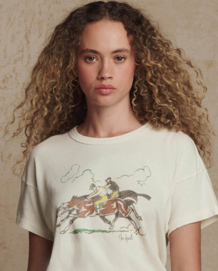 A woman with curly hair wears THE GREAT. PF26 The Boxy Crew w/ Derby Graphic in washed white, a vintage-inspired tee featuring two jockeys on horses, handcrafted in Los Angeles.
