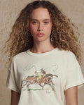 A woman with curly hair wears THE GREAT. PF26 The Boxy Crew w/ Derby Graphic in washed white, a vintage-inspired tee featuring two jockeys on horses, handcrafted in Los Angeles.