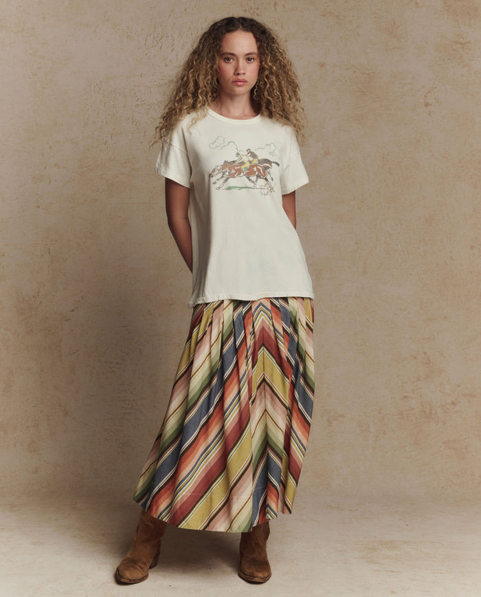 A person stands against a neutral background wearing THE GREAT. PF26 The Boxy Crew w/ Derby Graphic in Washed White, paired with a long multicolored striped skirt and brown boots—a handcrafted Los Angeles look.