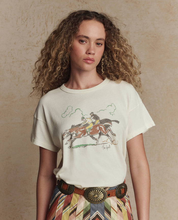 A woman with curly hair wears THE GREAT. PF26's The Boxy Crew w/ Derby Graphic in Washed White, pairing the vintage-inspired tee with patterned pants and a decorative belt for a laid-back Los Angeles look.