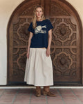 A woman stands before an ornate wooden door, wearing THE GREAT. PF26's The Boxy Crew w/ Everglade Graphic in True Navy, paired with a light skirt and brown boots—showcasing the effortless style of handcrafted Los Angeles fashion.