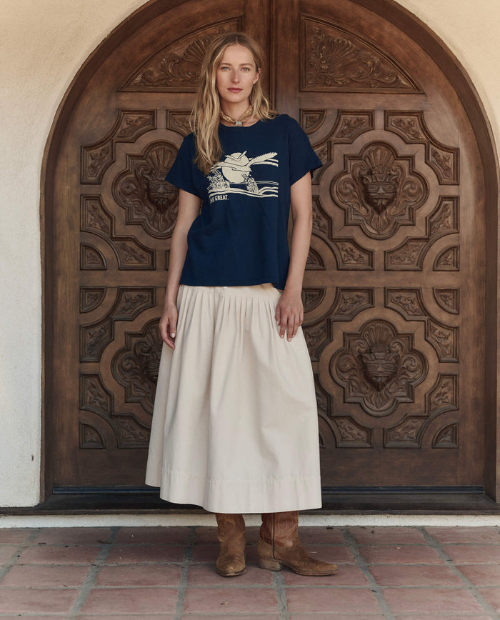A woman stands before an ornate wooden door, wearing THE GREAT. PF26's The Boxy Crew w/ Everglade Graphic in True Navy, paired with a light skirt and brown boots—showcasing the effortless style of handcrafted Los Angeles fashion.