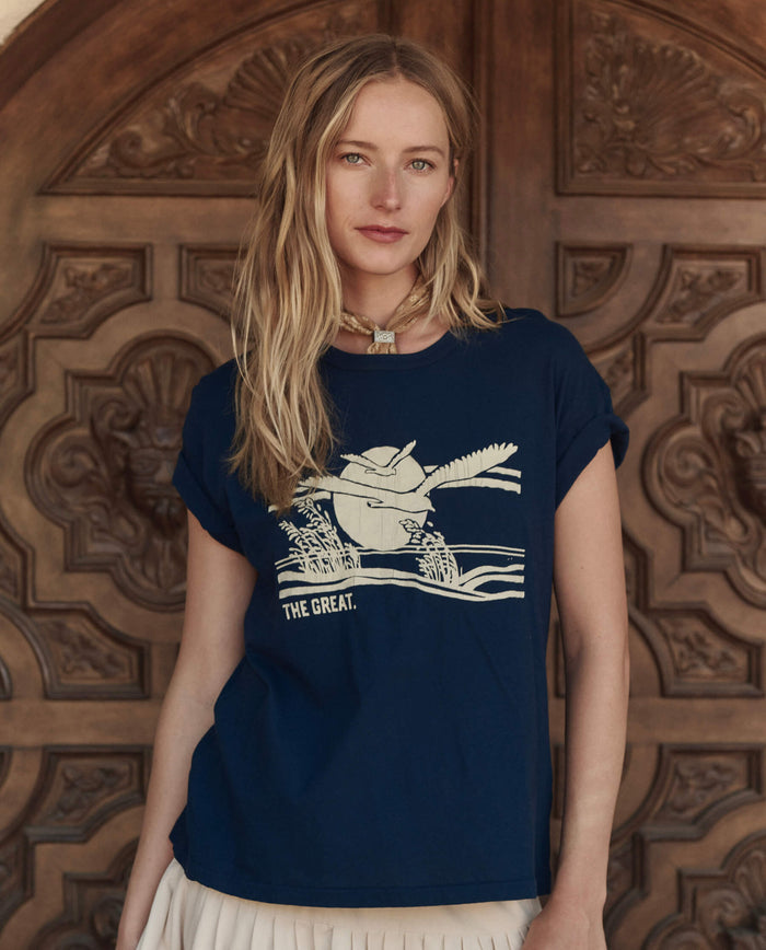 A woman with long blonde hair stands before a carved wooden door, wearing THE GREAT. PF26's The Boxy Crew w/ Everglade Graphic in True Navy and a light-colored skirt, all handcrafted in Los Angeles.