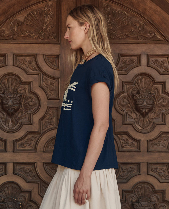 A woman with long blonde hair, wearing THE GREAT. PF26 The Boxy Crew w/ Everglade Graphic in True Navy and a cream skirt, stands in profile before an ornate wooden door.