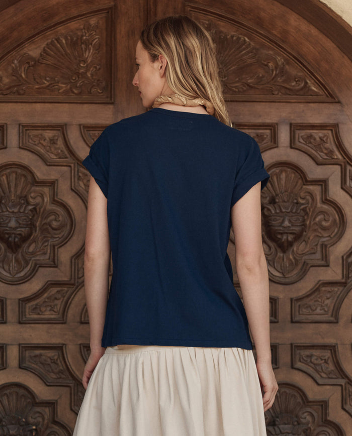 A woman with long blond hair faces away, wearing THE GREAT. PF26 The Boxy Crew w/ Everglade Graphic in True Navy and a light skirt, standing before an ornate wooden door.
