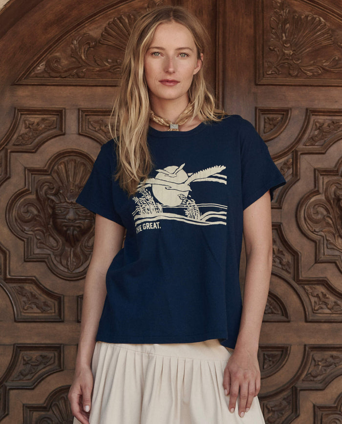 A woman with long blonde hair wears THE GREAT. PF26 The Boxy Crew w/ Everglade Graphic in True Navy and a cream skirt, standing before an ornate wooden door, exuding effortless handcrafted Los Angeles style.
