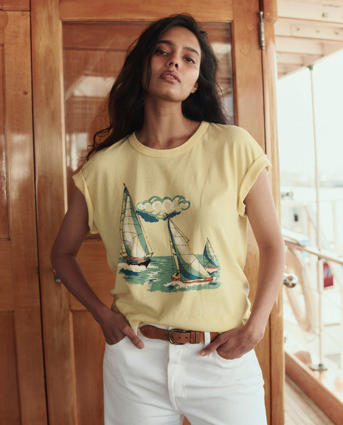 A woman stands indoors on a boat, wearing THE GREAT. SU26 The Boxy Crew W/ Sailing Boats Graphic in Sunbeam and white pants, hands in pockets, looking at the camera.