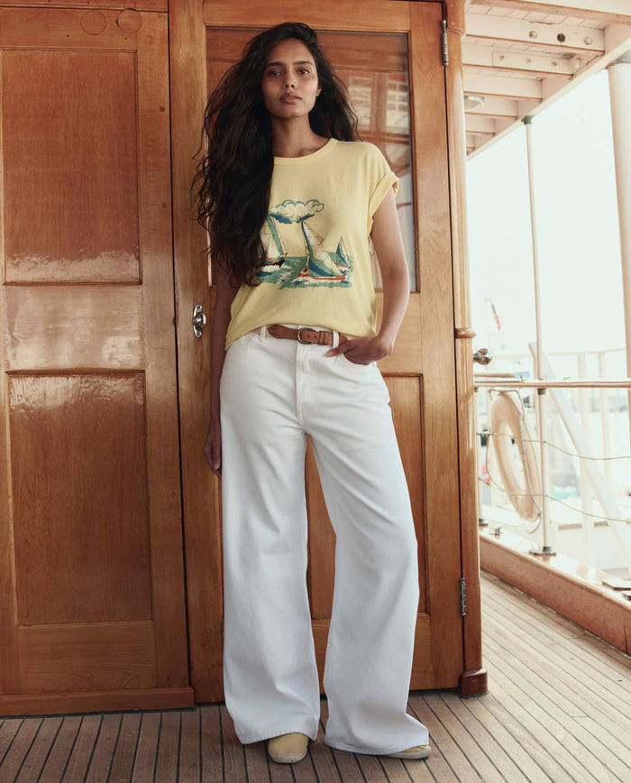 A woman stands on a wooden deck in front of a closed door, wearing THE GREAT. SU26’s The Boxy Crew W/ Sailing Boats Graphic in Sunbeam, styled with white wide-leg pants and light-colored shoes.