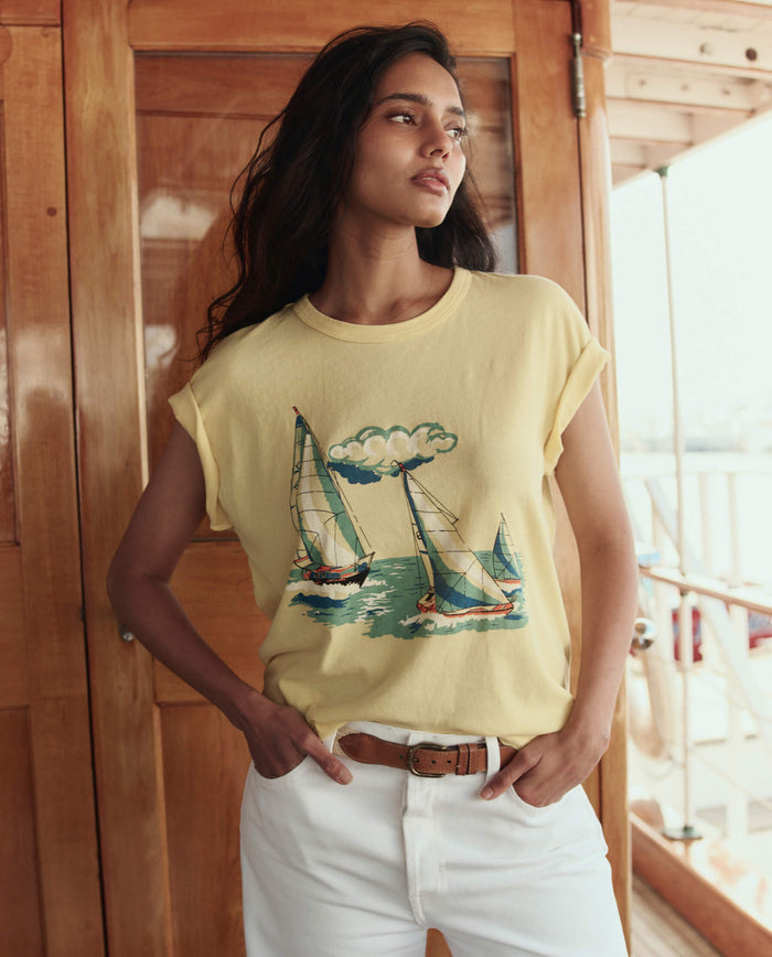 A woman stands indoors on a boat, wearing THE GREAT. SU26 The Boxy Crew W/ Sailing Boats Graphic in Sunbeam and white pants, with one hand in her pocket.