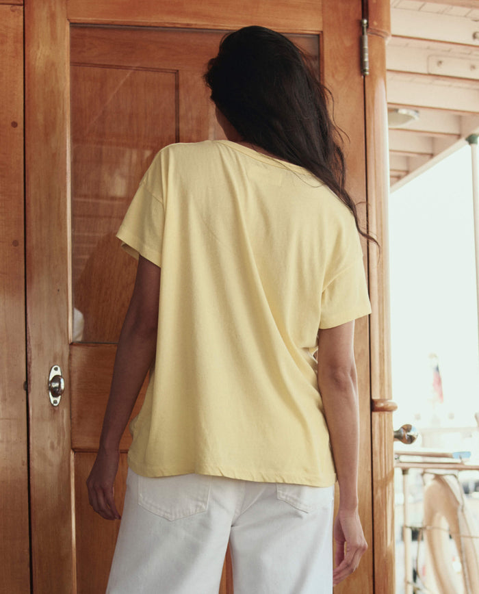 A person with long dark hair, wearing THE GREAT. SU26 The Boxy Crew W/ Sailing Boats Graphic in Sunbeam and white pants, stands with their back to the camera in front of a wooden door.