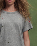 A person with curly hair wears THE GREAT. SU26 The Boxy Crew w/ Tossed Floral Embroidery in Heather Grey, standing in front of a green background.