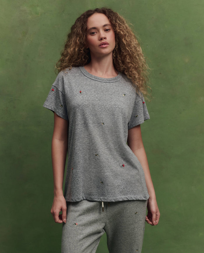 A woman with curly hair wears THE GREAT. SU26’s The Boxy Crew w/ Tossed Floral Embroidery in heather grey and matching sweatpants, standing against a muted green background.