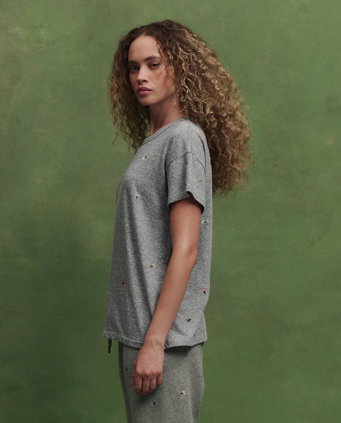 A person with curly hair stands in profile wearing THE GREAT. SU26 The Boxy Crew W/ Tossed Floral Emb. in Heather Grey against a green background.