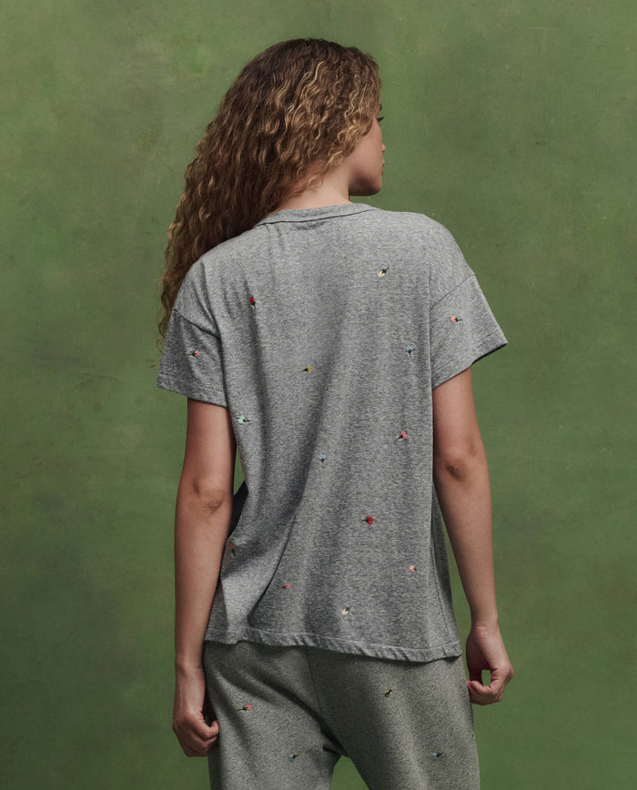 A person with long curly hair faces away, wearing THE GREAT. SU26's Boxy Crew W/ Tossed Floral Emb. in Heather Grey and pants with small colorful embroidered dots, set against a plain green background.