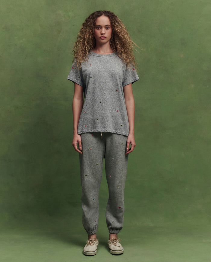 A woman faces forward against a green background, wearing THE GREAT. SU26's The Boxy Crew W/ Tossed Floral Emb. in Heather Grey with matching sweatpants and beige sneakers.