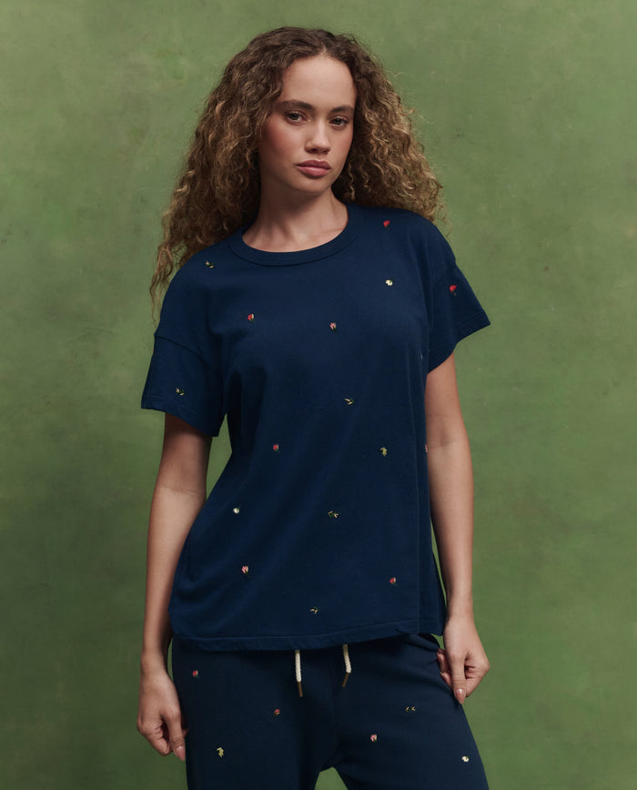 A woman with curly hair wears THE GREAT. SU26 The Boxy Crew W/ Tossed Floral Emb. in navy and matching pants, standing before a green background.