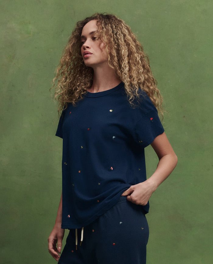 A woman with curly hair wears THE GREAT. SU26's The Boxy Crew W/ Tossed Floral Emb. in navy, paired with matching pants, as she stands against a green background.