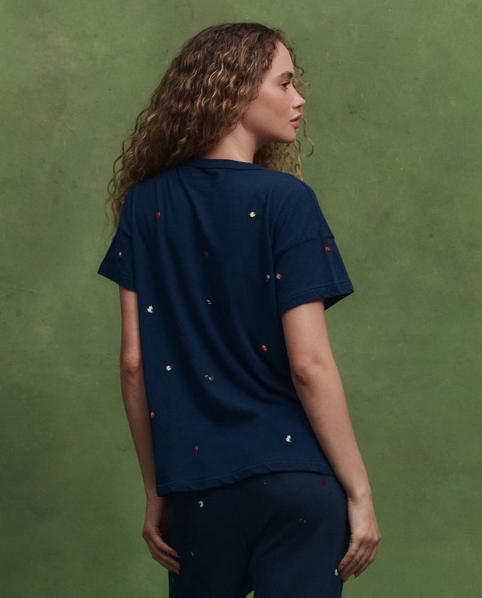 A person with curly hair, seen from behind, wears THE GREAT. SU26's The Boxy Crew W/ Tossed Floral Emb. in navy with floral embroidery, paired with matching pants, set against a green background.