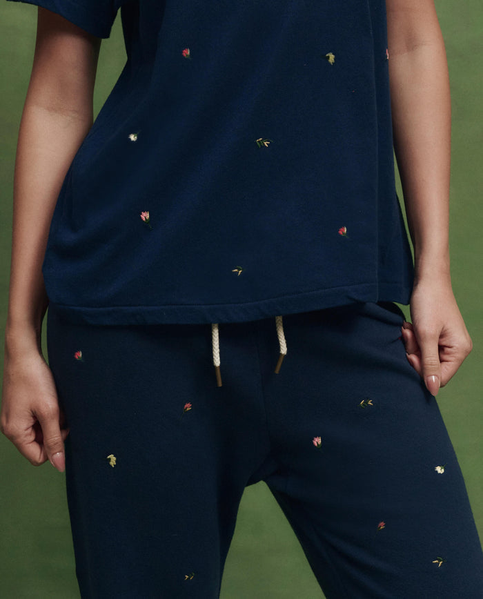 A person stands against a green background wearing THE GREAT. SU26 The Boxy Crew w/ Tossed Floral Embroidery in navy—boxy fit, small embroidered florals, and white drawstrings for a handcrafted Los Angeles vibe.
