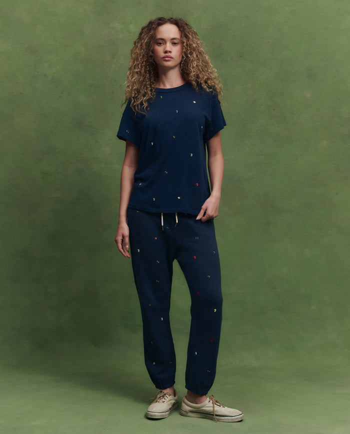 A person with curly hair wears THE GREAT. SU26 The Boxy Crew W/ Tossed Floral Emb. in navy and matching joggers with small colored dots, standing against a green background.