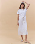 A woman stands against a beige background wearing THE GREAT. PF25 HEAVY JERSEY KNITS The Boxy Tee Dress in True White, styled with a cotton rope belt, white sandals, and a headband—effortless and made in Los Angeles.