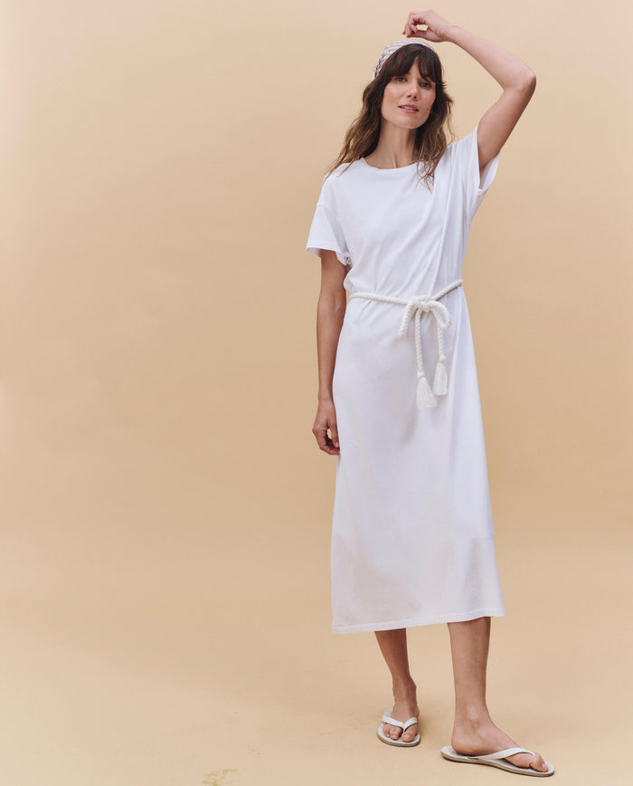 A woman stands against a beige background wearing THE GREAT. PF25 HEAVY JERSEY KNITS The Boxy Tee Dress in True White, styled with a cotton rope belt, white sandals, and a headband—effortless and made in Los Angeles.
