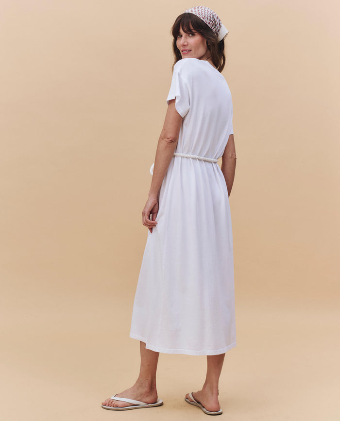 A woman stands sideways against a beige background wearing THE GREAT. PF25 HEAVY JERSEY KNITS The Boxy Tee Dress in True White, styled with a cotton rope belt, white flip-flops, and a patterned headscarf.