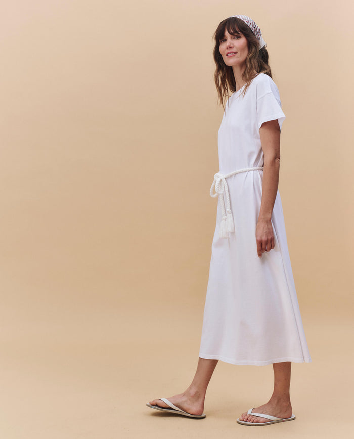 A person stands sideways against a beige background, wearing THE GREAT. PF25 HEAVY JERSEY KNITS The Boxy Tee Dress in True White, styled with a cotton rope belt, white sandals, and a patterned headband.