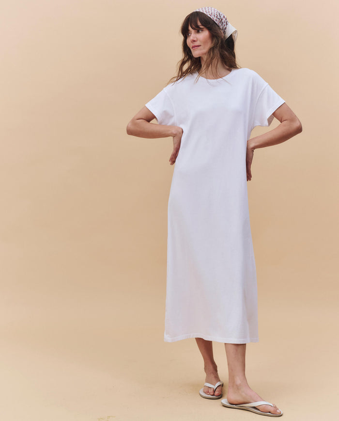 A woman stands against a beige background wearing THE GREAT. PF25 HEAVY JERSEY KNITS The Boxy Tee Dress in True White, styled with a white patterned headscarf and white flip-flops.