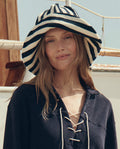 A woman stands outdoors near a boat, wearing THE GREAT. SU26 The Bucket Hat in Navy Shoreside Stripe and a navy lace-up top.