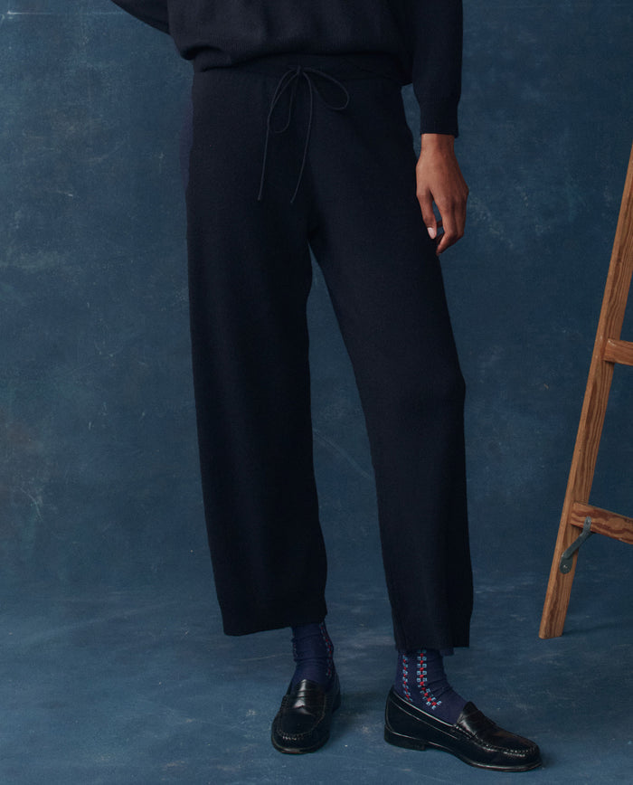A person wearing THE GREAT. H25 CASHMERE The Cashmere Lantern Pant in navy stands next to a wooden ladder against a dark blue textured backdrop, with only the lower half of their body visible.