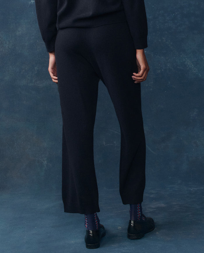 Person stands with their back to the camera in THE GREAT. H25 CASHMERE The Cashmere Lantern Pant in Navy, paired with a black top, patterned socks, and black shoes against a dark blue background—a refined luxury loungewear look.