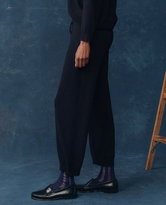 A person stands in front of a blue background wearing THE GREAT. H25 CASHMERE The Cashmere Lantern Pant in Navy, paired with black loafers and patterned blue socks; a wooden step ladder appears partially on the right.