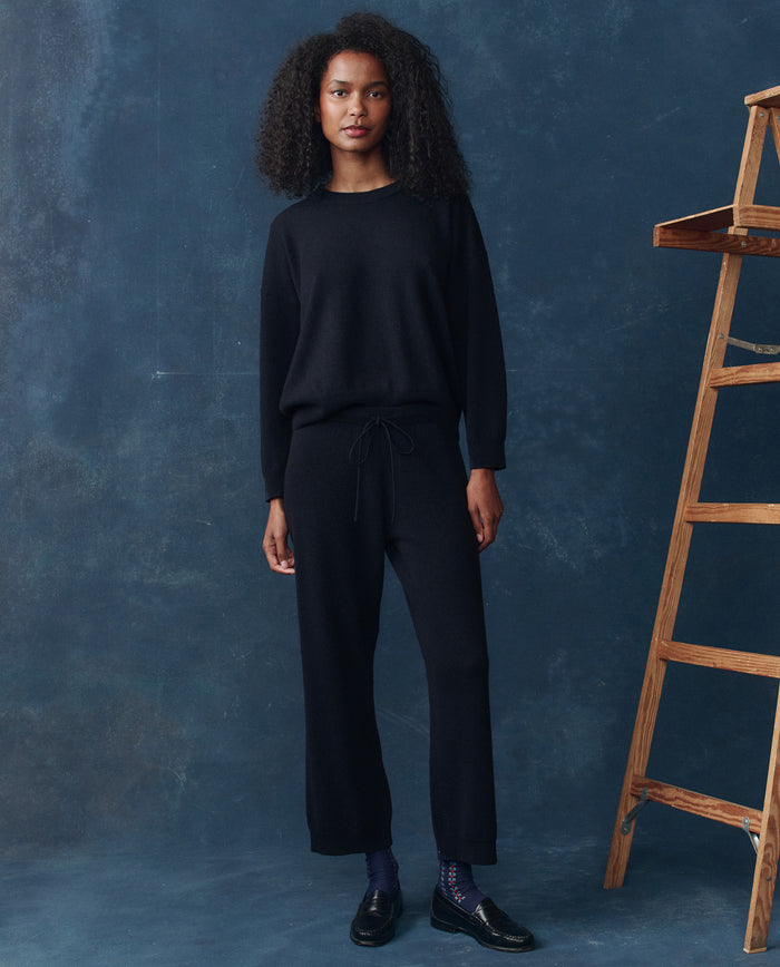 A person stands indoors wearing a black long-sleeve sweater, The Cashmere Lantern Pant in navy by THE GREAT. H25 CASHMERE, black shoes, and blue socks beside a wooden ladder against a dark blue background.