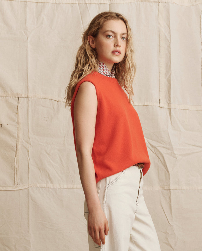 A person with wavy blonde hair wears THE GREAT. PS26's The Cashmere Pullover Vest in Hot Orange over a patterned shirt and white pants, standing before a beige fabric backdrop.