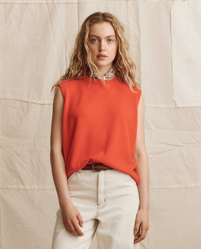 A person with wavy blonde hair wears THE GREAT. PS26 The Cashmere Pullover Vest in Hot Orange and light-colored pants, standing against a beige fabric backdrop.