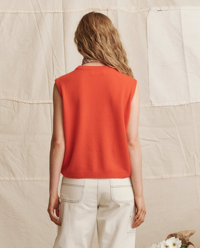 A person with wavy, light brown hair is seen from behind in THE GREAT. PS26 The Cashmere Pullover Vest in Hot Orange, paired with white jeans, standing before a beige fabric backdrop.