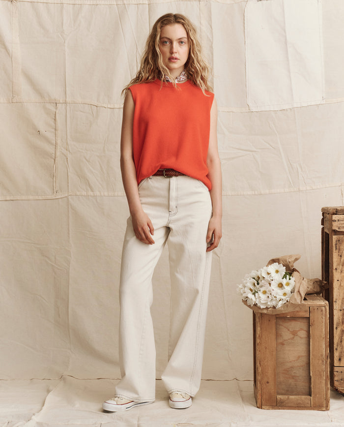 A woman stands indoors wearing THE GREAT. PS26 The Cashmere Pullover Vest in Hot Orange, paired with white wide-leg pants and shoes beside wooden crates and white flowers—her look radiates crisp, relaxed elegance with vintage menswear charm.