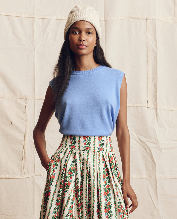 A woman stands before a neutral backdrop wearing THE GREAT. PS26's The Cashmere Pullover Vest in Soft Blue, styled with a beige knit beanie and a high-waisted floral skirt.