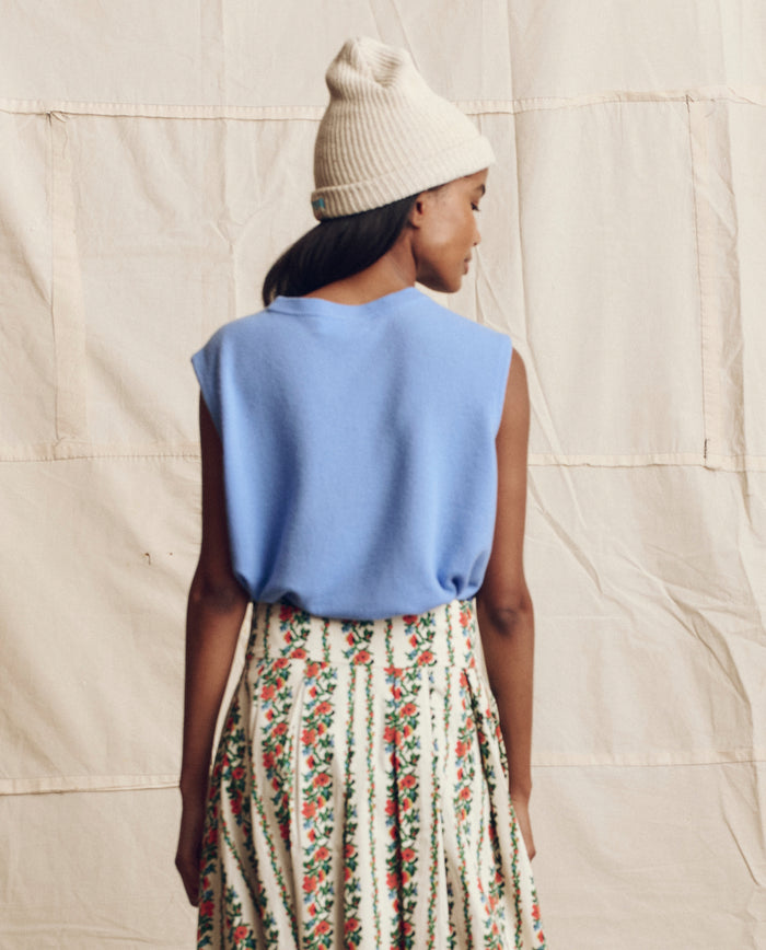A person stands facing away, wearing a cream knit beanie and THE GREAT. PS26 The Cashmere Pullover Vest in Soft Blue, paired with a floral skirt, set against a beige fabric backdrop.