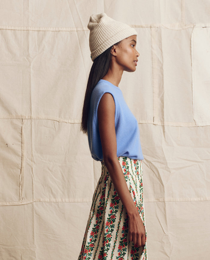 A woman in profile wears THE GREAT. PS26 Cashmere Pullover Vest in Soft Blue with a cream knit beanie and a white skirt featuring a red and green floral pattern, standing against a beige fabric backdrop.