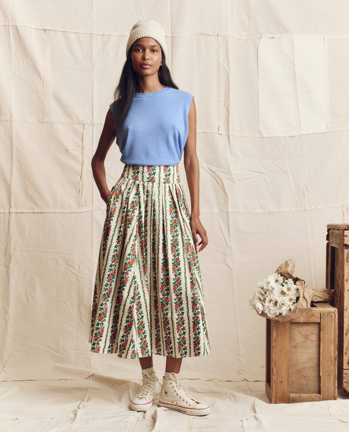 A woman stands indoors in THE GREAT. PS26 The Cashmere Pullover Vest in Soft Blue, with a floral and striped midi skirt, white beanie, and high-top sneakers beside wooden crates and white flowers for vintage menswear charm.