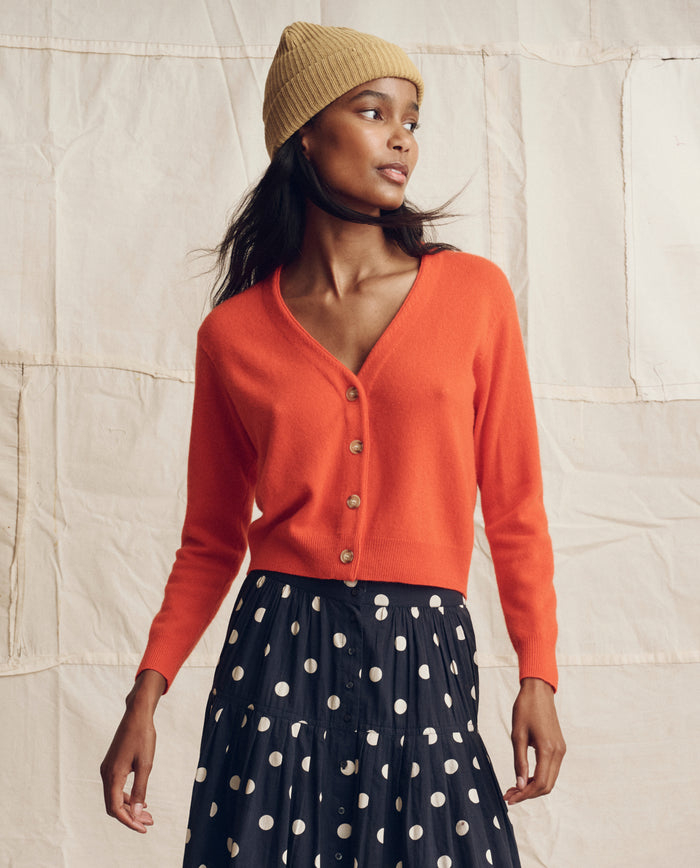 A woman wearing THE GREAT. PS26 The Cashmere Tiny V Neck Cardigan in Hot Orange, a tan knit beanie, and a black skirt with white polka dots stands before a beige fabric backdrop.