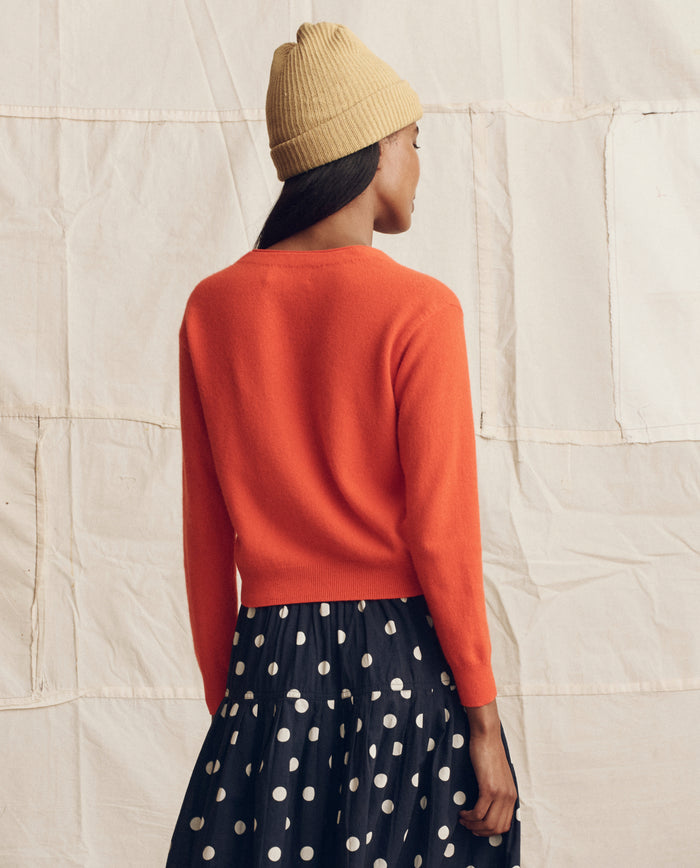 A person stands facing away against a neutral fabric backdrop, wearing THE GREAT. PS26 The Cashmere Tiny V Neck Cardigan in Hot Orange, a beige knit hat, and a black skirt with white polka dots.
