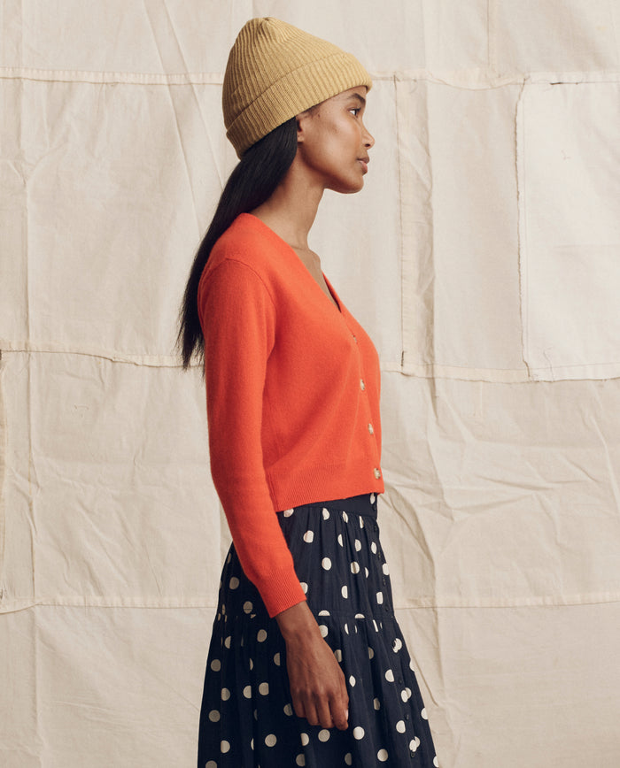 A woman in profile wears THE GREAT. PS26 The Cashmere Tiny V Neck Cardigan in Hot Orange over a navy polka dot skirt, standing against a beige quilted fabric backdrop and accessorized with a yellow knit beanie.