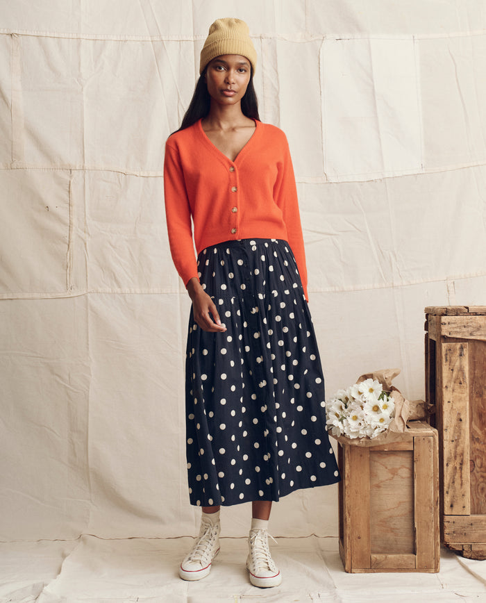 A woman in THE GREAT. PS26 The Cashmere Tiny V Neck Cardigan in Hot Orange stands indoors beside wooden crates and a bouquet of flowers, wearing a beige beanie, black polka dot skirt, and white sneakers.