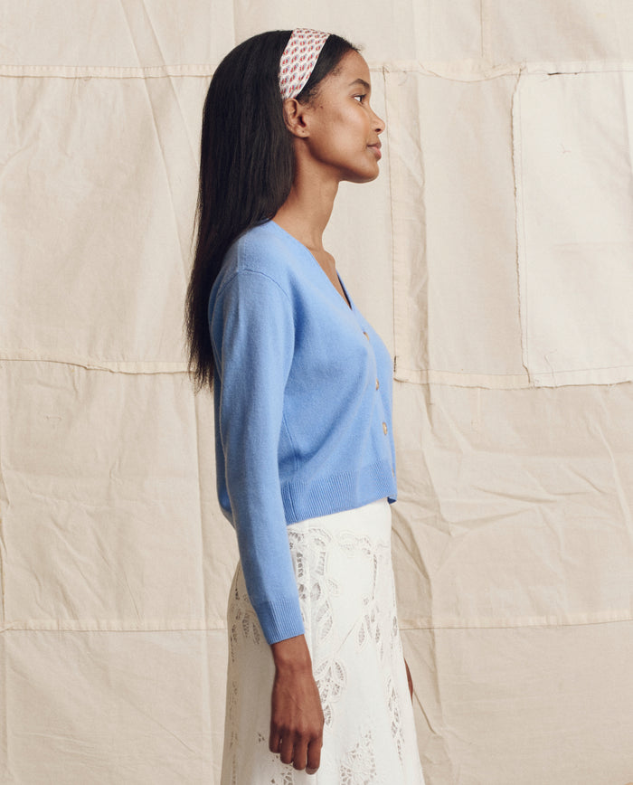 A woman stands in profile against a beige fabric backdrop, wearing THE GREAT. PS26 The Cashmere Tiny V Neck Cardigan in Soft Blue, paired with a white lace skirt and a pink patterned headband.