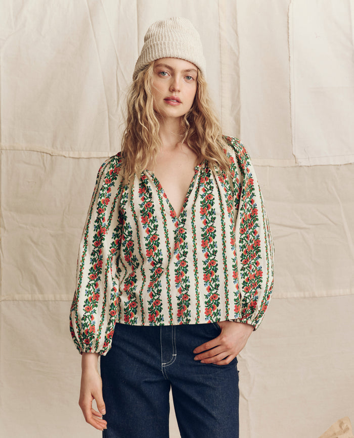 A person wearing THE GREAT. PS26 The Chariot Top in Tapestry Floral with an open neckline, balloon sleeves, a cream knit beanie, and dark jeans stands before a beige fabric backdrop.